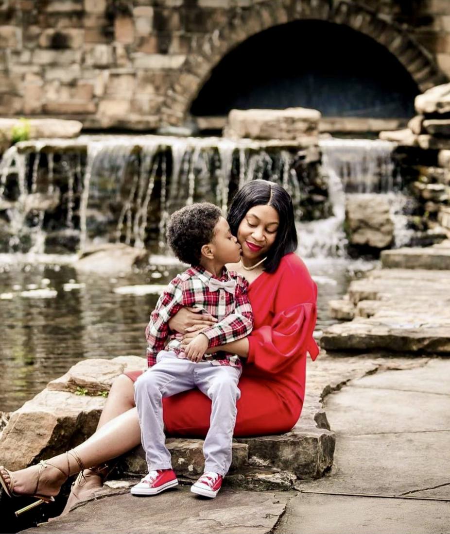Raven in a red dress holding her son Khaza in front of a stone waterfall as he kisses her cheek.